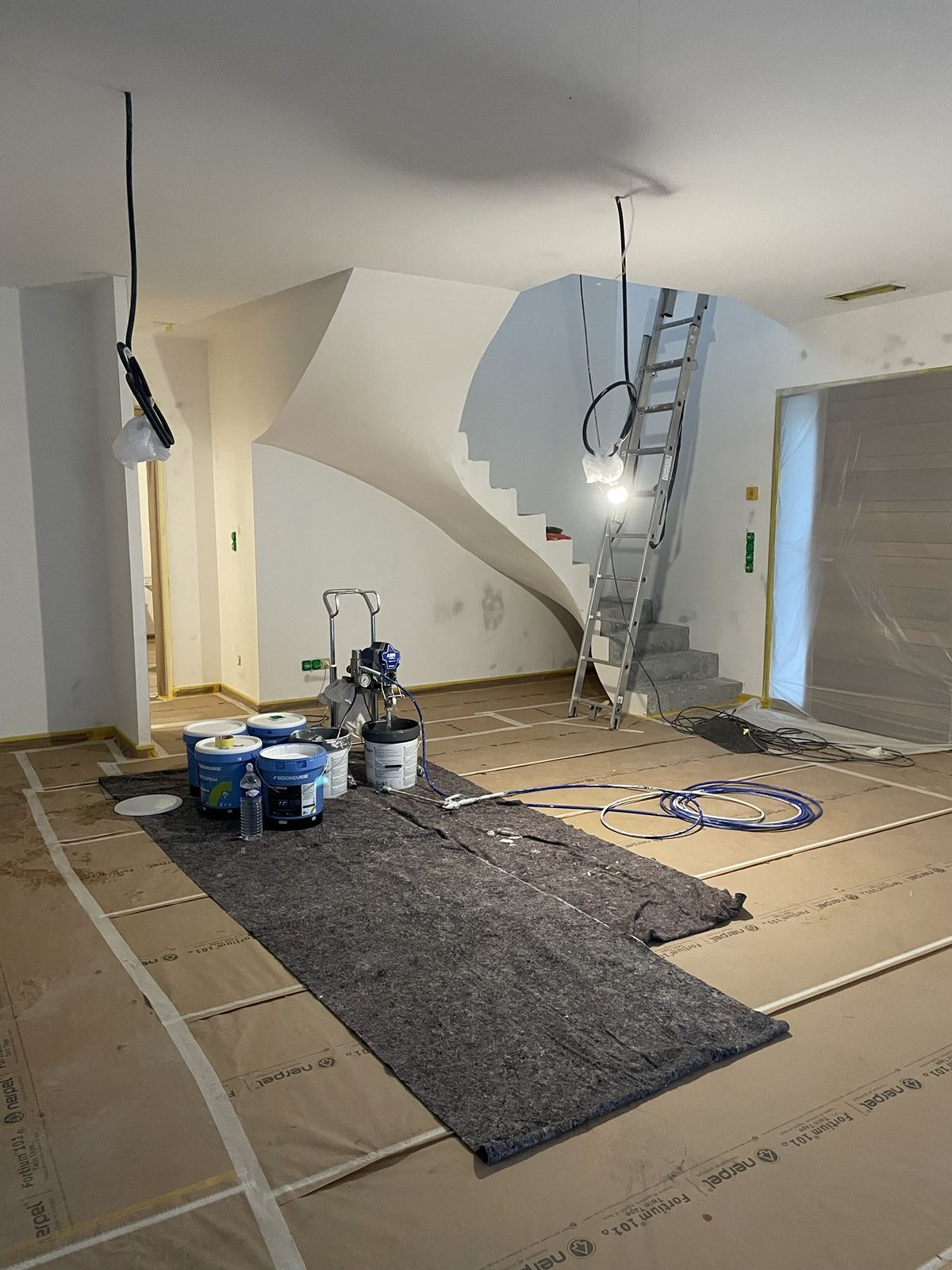 Pistolet airless et pots de peinture au centre d’un séjour protégé, escalier en arrière-plan